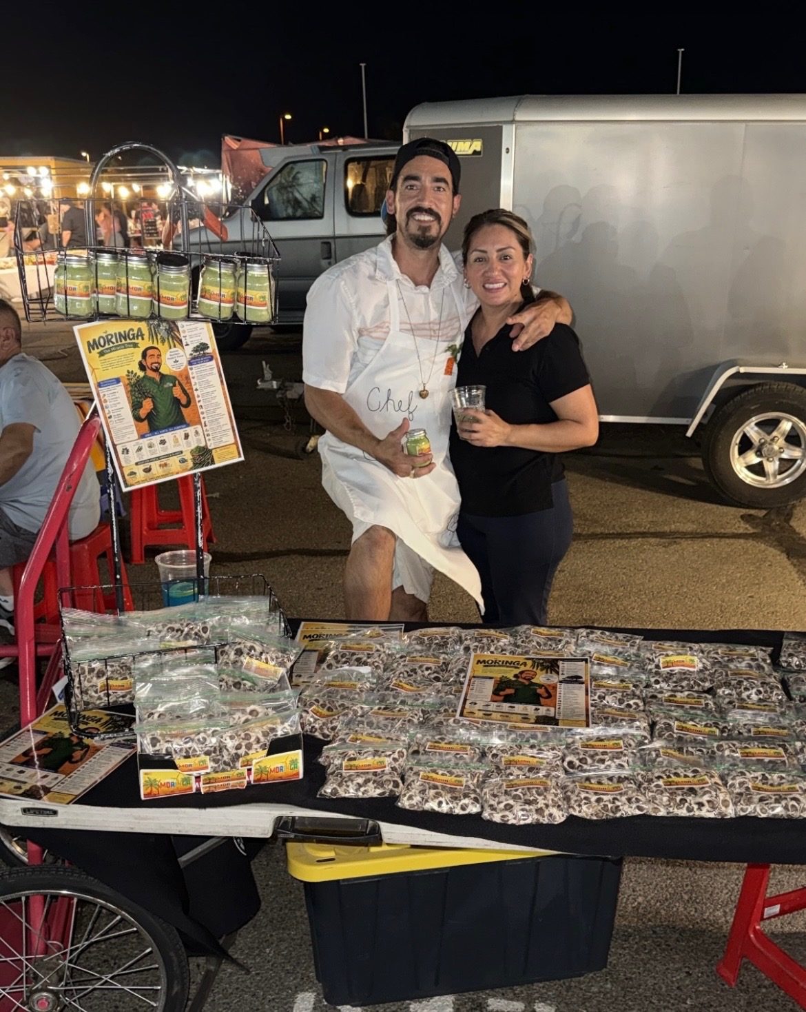 JP sharing a side hug with his girlfriend at his Moringa stand at the Indio swap meet, product and brand signage on the table in front of them